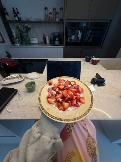 In a modern kitchen, a plate of sliced strawberries dusted with powdered sugar sits on a marble countertop. Next to the plate are white earbuds connected to a black notebook, a small green cup, and an ornate rectangular dish. Further back, a laptop is open, and a small black video camera rests on a white square. Two small bowls, one dark blue and one brown, are positioned on the edge of the counter. In the background, shelves display kitchenware, including bottles of wine, jars, and a coffee machine. The kitchen appears to be located in a home in Ridderkerk, Netherlands.