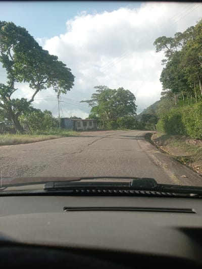 The image depicts a view from inside a vehicle, looking out onto a paved road winding through a rural area. The foreground shows the dashboard and windshield wipers of the vehicle. The road ahead is cracked and shows signs of wear. On either side of the road are trees and vegetation, with a small, run-down building visible on the left side of the road. In the distance, a mountain can be seen. The sky is partly cloudy. The image is taken in Honda, Venezuela.