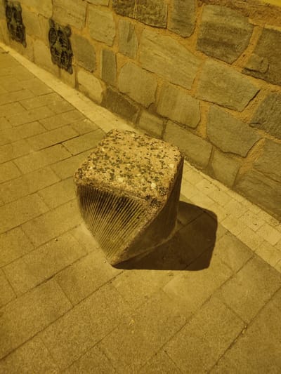 A close-up, eye-level shot captures a roughly cubical, speckled concrete bollard positioned on a paved sidewalk. The bollard, with its rough, textured top surface, is tilted, with one side exhibiting deep, vertical grooves. Its shadow is cast to the right, across the paving stones. Behind the bollard, a textured stone wall runs horizontally, composed of irregularly shaped, muted gray stones interspersed with a yellowish mortar. Two dark, ornate decorative elements, possibly metalwork, are embedded in the wall to the left of the frame. The overall lighting suggests an evening or nighttime scene, with a warm, yellowish hue illuminating the environment. The image does not contain any people or indicate any specific actions or interactions. The setting appears to be an outdoor urban environment. Without further information, it is not possible to definitively identify the location within Osmangazi, Turkey.