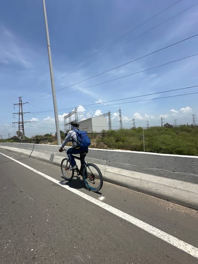 A person is riding a bicycle on a paved road, viewed from behind and slightly to the side. The rider is wearing a grey long-sleeved shirt, blue jeans, a helmet, and a blue backpack. The bicycle is a dark color with silver spokes and blue accents on the rims. The road has a white line marking the lane. In the background, there is a concrete barrier, a field of green bushes, and several electrical transmission towers and structures under a bright blue sky with scattered clouds. The scene appears to be in an open, possibly semi-industrial area. This location is identifiable as the area near the Barranquilla substation and the connected highway on the outskirts of Barranquilla, Colombia, based on the distinctive electrical infrastructure and surrounding landscape.