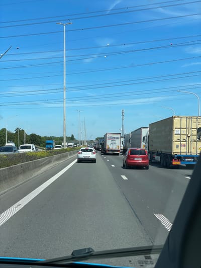This image shows a traffic jam on a highway. The perspective is from inside a vehicle, likely a car or truck, looking forward. The highway has multiple lanes, and several vehicles are visible, including trucks, a red car, and a white car. The scene is set outdoors with a clear blue sky. Tall, white poles with curved tops line the side of the highway, and overhead power lines stretch across the frame. In the distance, more vehicles and what appear to be wind turbines can be seen. The overall mood is one of congestion and potentially frustration, common during highway traffic jams.
There is some visible text on the vehicles. The large truck in the foreground has text that includes "SINGTRANS". Another truck further back has text like "SKYU 80670" and "2 2823 JXM". The red car has a license plate that reads "2 ZSU 220". The white car has a license plate with "2 BXX 228". The time of day appears to be daytime, given the bright sunlight and clear sky, and the weather seems fair. The location context provided is Temse, Belgium, which is consistent with the types of vehicles and infrastructure seen. Some small, spherical objects, possibly balloons, are visible suspended from the power lines against the blue sky.