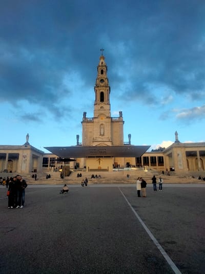 The image depicts the Sanctuary of Fátima in Fátima, Portugal, at dusk. The central subject is the Basilica of Our Lady of the Rosary of Fátima, a prominent light-colored stone building with a tall, ornate bell tower topped by a cross, visible under a partially cloudy, blue-grey sky. In front of the Basilica, a large, modern, flat-roofed canopy structure extends over an elevated area accessed by wide steps. A large crucifix is visible under this canopy. To the left and right, grand, colonnaded structures, consistent with the sanctuary complex, flank the main basilica. The foreground is a vast, paved esplanade with white demarcation lines.
Numerous individuals are scattered across the scene. In the immediate foreground on the left, three people (two adults and one child) stand together, one adult holding a phone. On the right foreground, two adults and a child are observed standing. A single individual is crouched near a small wheeled object in the mid-ground. Many other people are seated or standing on the steps leading to the main structures and under the canopy, appearing to be visitors or pilgrims. Artificial lights are visible under the canopy and within parts of the surrounding buildings. The overall scene suggests a time of day transitioning from late afternoon to evening, with a moderate number of people present in the sacred space.