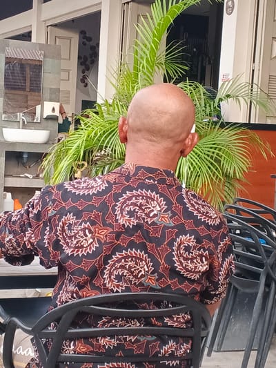 The image shows the back of a bald man wearing a dark-colored shirt with a repeating pattern of paisley-like motifs in shades of red, white, and turquoise. He is seated in a black metal chair, facing away from the camera. Behind him, a lush green palm plant is visible, with its fronds extending upwards and outwards. In the background, there appears to be a building with white window frames and a glimpse of an open doorway. To the left, a sink and a mirror mounted on a gray tiled surface are partially visible, suggesting an outdoor or semi-outdoor wash area. Stacked black metal chairs are to the right of the man. The scene is set in what appears to be an outdoor seating area, possibly at a cafe or restaurant, in Geulumpangsulu Timu, Indonesia.