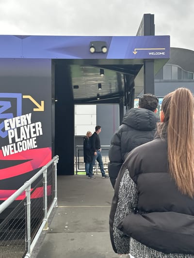 The image captures a scene at a venue in Dartford, United Kingdom. The foreground features a blue and black sign with "WELCOME" printed vertically, and "EVERY PLAYER WELCOME" displayed prominently, along with arrow graphics. A metal barricade is visible near the bottom of the frame. Further back, there are people walking through an entryway, including a couple and other individuals in dark clothing. One person in the foreground is wearing a thick black jacket. The architectural design of the building is partially visible in the background. The weather appears overcast, with a gray sky.