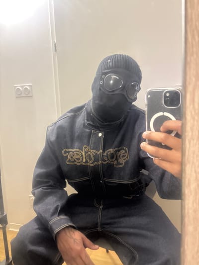 The image depicts an indoor scene in Besançon, France. A lone individual is captured in a mirror selfie, appearing seated. The subject wears a dark blue denim ensemble, possibly a jumpsuit, featuring prominent gold contrast stitching and a large, mirrored script logo embroidered in gold thread across the chest, which reads "agaisneleB" (Balenciaga in reverse). A black ribbed beanie covers the head, and a black balaclava obscures the lower face and neck, extending to cover the upper forehead. Dark-tinted goggles, with visible side vents or hardware, cover the eyes. The subject's reflected right hand holds a silver-colored smartphone, consistent with an iPhone, housed in a clear case with a visible white circular MagSafe ring. The reflected left hand rests on the upper thigh, revealing a dark-colored tattoo on the inner forearm. The background consists of a light-colored wall with a white double light switch or outlet plate, one indicator light illuminated in orange. A narrow, light-colored wooden frame, likely a doorframe or mirror frame, runs vertically along the right side of the image. A black or dark-colored metal stand is partially visible in the lower left corner. The floor beneath the subject is light-colored wood.