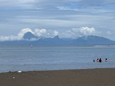 The image depicts a coastal scene in Māhina, French Polynesia, France. The foreground consists of a dark-colored sand beach with minor light-colored debris. The midground features calm, dark blue-grey ocean water, where four individuals are partially submerged on the right side, appearing to be wading or swimming. To the left of center in the midground, a white buoy with a vertical pole is present. The background displays a distant, mountainous landmass, primarily dark green, with several peaks partially obscured by large white cumulus clouds. The sky above is mostly overcast with scattered grey and white clouds.