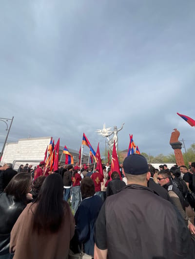 A gathering of people is shown at a monument in Akhuryan, Armenia. The monument features a statue of a woman with a sunburst behind her. Many individuals are holding Armenian flags. The monument is surrounded by people, and there is a stone structure visible in the background. The sky is overcast.