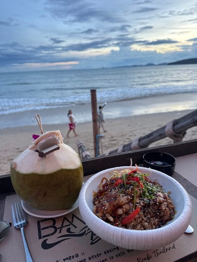 The image shows a close-up of a meal and a drink on a table at a beachside restaurant. In the foreground, there is a whole coconut with a straw and a garnish of what appears to be white bread and a purple flower. To the right of the coconut is a bowl of food, which appears to be rice topped with chicken in a sauce, garnished with chopped scallions, red chili slices, and sesame seeds. A fork rests to the left of the coconut, and a pair of sunglasses are visible on the far left edge of the table. In the background, the ocean and beach are visible. Two people are walking on the beach. The sky is cloudy with hints of pink and orange, suggesting it is either sunrise or sunset. The restaurant appears to be located on a beach in Ban Klong Haeng, Krabi, Thailand, as indicated by text on the napkin in the foreground.