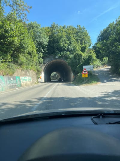 The image shows a road leading to a tunnel entrance in Kruče - Kërruçi, Montenegro. The road is asphalt, with white lane markings. On the left side of the road, there is a concrete wall with graffiti on it. The tunnel is made of stone and appears to be old. There are trees and vegetation on either side of the road and surrounding the tunnel. A traffic sign is visible on the right side of the road, indicating a tunnel ahead and a distance of 93 meters to Kruče. The sky is blue and clear. The perspective is from inside a vehicle, likely a car, with the dashboard visible in the foreground. Two vehicles are seen approaching the tunnel, one appears to be a motorcycle.
