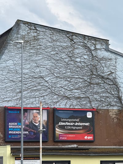 The image shows the side of a building in Moers, Germany, covered in ivy. Two billboards are mounted on the wall. The upper billboard advertises "E.ON Highspeed" internet services, featuring a graphic of a swirling vortex and the E.ON logo. Text on the billboard reads: "Leistungsstarkes Glasfaser-Internet: E.ON Highspeed" and "Für Ihr Zuhause: eon-highspeed.com it's on us". The lower billboard advertises "Diakonie Pflege" (Diaconia Care) and features a portrait of an elderly man. The text on this billboard includes: "AUCH DU BRAUCH T PFLEGE. IRGEND ANN.", "MACH DICH #Stark", and "pflege.diakonie.de". A third, partially visible billboard on the right seems to be for "RECKMANN AUTOMOBILE Moers". The sky appears overcast, suggesting a cloudy day. Metal poles and fasteners are visible, indicating the billboards are mounted outdoors.