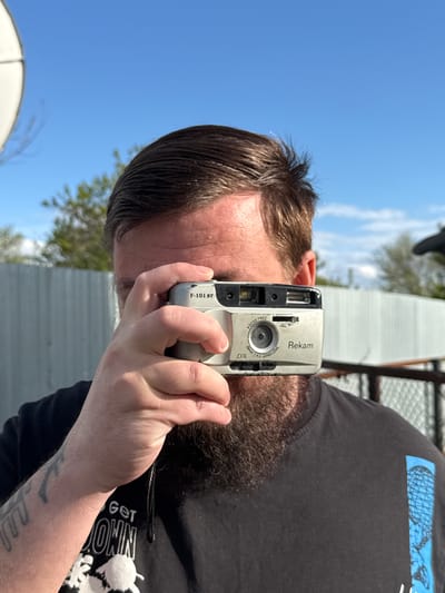 An outdoor, eye-level shot captures a person, likely male, in Aksay, Kazakhstan, holding a compact film camera up to their eye. The person's right hand grips the camera, obscuring most of their face except for their dark hair, forehead, and part of a beard. They appear to be in the process of taking or framing a photograph.

The camera is silver and black with various details visible. Text on the camera includes "F-101 ST", "FOCUS FREE", "25mm 1:4.5 JAPAN LENS", "Rekam", and "DX". The person is wearing a black t-shirt, on which partial white text "GET ONIN" (likely part of a larger phrase) and some white graphic elements are visible. A faint tattoo can be seen on their forearm.

The scene is set outdoors under a bright, clear blue sky with a few wispy clouds, indicating daytime. In the background, a light-colored building with a corrugated texture is visible, along with some green tree foliage. A large, curved white object, possibly a satellite dish, appears in the lower left portion of the frame.