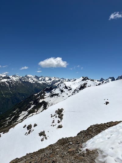 The media file depicts a vast mountain landscape under a clear blue sky. The primary subject is the rugged mountain range, characterized by steep slopes and jagged peaks. Patches of snow are scattered across the mountains, contrasting with the dark rock and green vegetation on the lower slopes. The scene suggests an outdoor environment, likely a natural park or remote wilderness area, given the lack of man-made structures.
The composition of the image implies a sense of scale and grandeur, with the mountains stretching into the distance. The bright sunlight indicates it is daytime, and the clear sky suggests fair weather. The presence of snow implies a high altitude or a season with cold temperatures, likely summer in a high mountain region. The location context provided, "Верхне-Тебердинское сельское поселение, Russia," points to the area being in the Karachay-Cherkess Republic of Russia, known for its mountainous terrain and natural beauty.
There are no visible people or objects in the image, and no discernible activity or event is taking place. The focus is solely on the natural landscape. Notable details include the sharp contrast between the snow-covered and rocky parts of the mountains, the distinct layering of the terrain from green slopes to snow-capped peaks, and the small, scattered clouds in the expansive blue sky. The overall impression is one of serene isolation and the imposing beauty of nature. No text is visible in the media file.