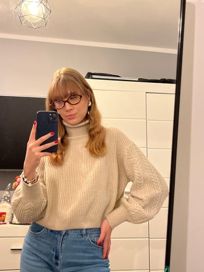 A young woman poses for a selfie in a mirror. She wears round glasses, a cream-colored turtleneck sweater with cable-knit sleeves, and blue jeans. Her blonde hair is styled with bangs. She holds a dark-colored smartphone in her right hand. A light fixture with a geometric design hangs from the ceiling. The reflection includes a white wardrobe and other room elements.