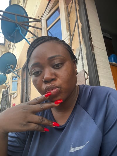 A medium close-up angled shot shows a person with dark skin and braided hair. They are wearing a dark blue t-shirt featuring a Nike swoosh logo on the upper right. Their nails are long and painted red. They have a nose ring on their left nostril. The person is looking at the camera, and their hand is near their face.
In the background, there is an overhang with a glass roof supported by metal beams. Also visible are multiple blue satellite dishes mounted on metal frames. The location appears to be an outdoor setting, possibly a rooftop in Dushepe, Nigeria.