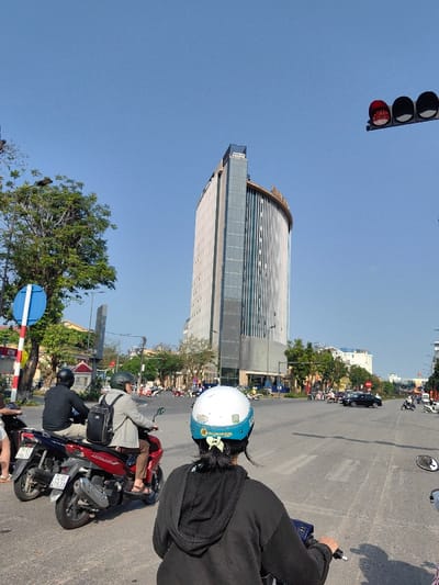 The image depicts a street scene in Thuận Hóa Ward, Huế City, Vietnam, on a clear, sunny day. In the foreground, viewed from behind, is a person wearing a white and light blue helmet with a yellow ribbon tied in their dark hair, seated on a dark-colored motorbike.
Directly in front of this person, slightly to the left, are two other motorbikes. The closest is a red scooter with two occupants; the rider wears a dark green helmet and a light grey long-sleeved shirt with a black backpack, while the passenger wears a dark long-sleeved shirt. Behind the red scooter is a dark blue motorbike with a single rider wearing a black helmet and a black long-sleeved shirt. All individuals on motorbikes are stationary, facing away from the viewer.
To the left, a blue and white circular road sign on a red and white striped pole is visible, partially obscured by green foliage from a large tree. An ATM sign is also visible on the pole. Above the street on the far right, a set of traffic lights displays a solid red signal.
Dominating the background and center-right of the image is a tall, modern building, identified as Vinpearl Hotel Huế, featuring a curved glass facade with vertical grey and lighter panels, and a distinctive gold-colored architectural accent at its apex. The sky above is clear and bright blue. Further down the street, beyond the intersection, more motorbikes and a few cars are visible, indicating an active urban environment.