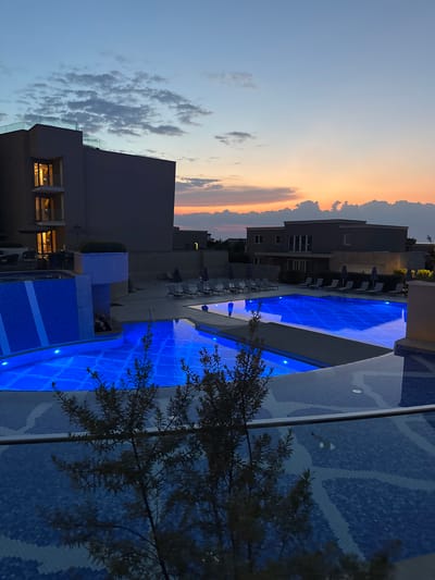 The image captures a serene evening scene at a resort in Crveni Vrh, Croatia. The main subject is a brilliantly lit swimming pool, its deep blue water illuminated by underwater lights, creating a vibrant and inviting atmosphere. The pool is lined with a mosaic tile pattern in shades of blue, adding to its visual appeal.

Adjacent to the pool, lounge chairs and umbrellas are neatly arranged, suggesting a relaxing day that has transitioned into evening. Several buildings with modern architecture are visible in the background. Some of these structures have illuminated windows, hinting at activity within. A notable detail is a tree with dark, feathery foliage in the foreground, its branches casting shadows on the illuminated pool.

The sky above is a canvas of soft twilight colors, transitioning from a pale blue near the horizon to warmer hues of orange and pink where the sun has set, indicating it is dusk. Scattered clouds add texture to the sky. The overall mood is tranquil and peaceful, evoking a sense of calm and relaxation, perfect for a vacation setting. There is no visible text in the image.