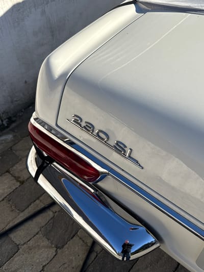 The image provides a close-up, angled view of the rear quarter panel of a white vintage car, likely a Mercedes-Benz 230 SL "Pagoda," indicated by the "230 SL" emblem clearly visible in chrome lettering on the trunk lid. The car's paintwork appears well-maintained, though some minor dust or water spots are visible on its surface.
The setting is outdoors in Strömstad, Sweden, under bright, direct sunlight, creating strong reflections and distinct shadows. A prominent chrome bumper curves around the rear of the car. Within its highly reflective surface, a partial reflection of the blue sky, the textured paved ground, and a person are visible. The person, likely the photographer, is wearing a dark jacket and appears to be holding a device, possibly a camera or phone.
A red rectangular taillight, framed by chrome trim, is present on the right side of the vehicle's rear. Small, indistinct markings or numbers can be faintly seen on the taillight lens. The ground reflected in the bumper and visible along the bottom edge of the frame is a paved surface, possibly cracked concrete or asphalt. The overall impression is of a classic car captured on a clear, sunny day.