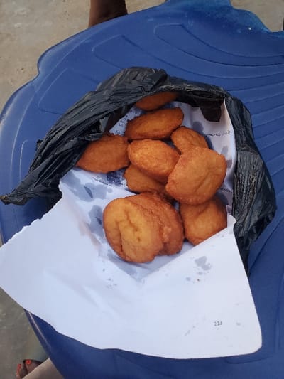 A black plastic bag, lined with white paper, holds approximately 10-15 golden-brown fried dough items. The white paper shows grease stains and has the number "213" printed in its lower right corner. The bag rests on a textured blue plastic surface, likely an outdoor chair or table. A dark-skinned arm is partially visible in the upper right, and a human foot with red-painted toenails is visible in the lower left. The setting is in Isunba, Nigeria.