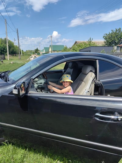 In Krestovo-Gorodishche, Russia, a young girl wearing a yellow bucket hat with dinosaur print and a pink and purple tank top sits behind the steering wheel of a black car, holding the wheel with both hands. She is smiling and looking towards the viewer. The car's interior is visible, with beige leather seats and a wooden steering wheel. The car is parked on a grassy area next to a dirt road, with houses and trees in the background under a bright blue sky with white clouds. Electrical poles and wires are also present in the background.