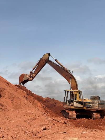 An excavator is actively digging into a large pile of reddish-brown soil under a partly cloudy sky. The excavator is yellow with some rust and dirt, and its bucket is filled with soil. The surrounding environment is an open area with mounds of earth, suggesting a construction or excavation site. The image depicts a typical scene in Jos, Nigeria, known for its earthmoving activities due to its topography and development projects.