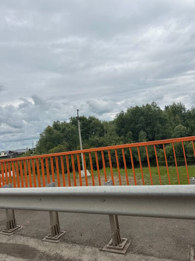 The image captures an overcast day in the village of Kuzhanovo, Russia, seen from what appears to be a bridge or overpass. In the foreground, a grey metal guardrail is prominently displayed, secured by sturdy metal supports anchored to the asphalt road surface. Behind the guardrail, an orange metal fence with vertical bars extends horizontally across the frame.
Beyond the fence, a lush green field is visible, dotted with patches of white flowers. Further in the background, a dense line of mature trees forms a natural barrier, their dark green foliage contrasting with the lighter sky. To the left, nestled amongst the trees, are several residential buildings with light-colored roofs, suggesting a small settlement or village. A utility pole with power lines stretches across the middle ground, adding to the rural infrastructure. The sky is filled with heavy, grey clouds, indicating a cloudy or possibly rainy weather condition. The overall scene depicts a tranquil, rural landscape under a somber sky.