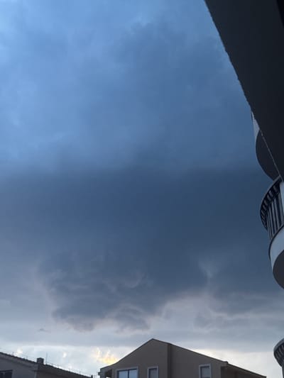 The image depicts a sky with dramatic cloud formations, possibly indicating an approaching storm or the aftermath of one. A significant portion of the frame is filled with dark, heavy clouds that create a somber and somewhat ominous atmosphere. To the right, a building with visible windows and balconies suggests an urban or suburban setting. The building is light-colored and appears to be modern in construction. The top of the image features a curved structure, possibly part of a building's architectural element or a balcony railing, with horizontal lines.
The lighting suggests it is either early morning or late afternoon, given the quality of light and the contrast between the dark clouds and the brighter areas. The overall weather appears unsettled, with a mix of dark and lighter clouds. The image is oriented vertically, with the sky occupying the majority of the frame. No people are visible in the image. The location context provided is "Trap, Montenegro," which hints at the geographical setting but offers no specific visual cues within the image itself. There is no visible text in the image.
