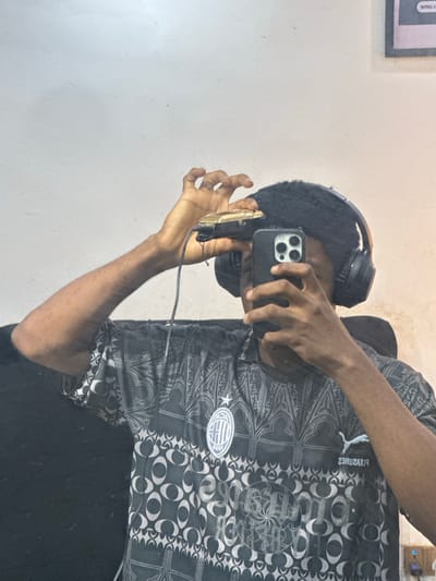 A person is standing indoors, holding a smartphone in their hands and appearing to take a photo or video. They are wearing headphones and a shirt with a distinctive pattern. The environment is sparsely decorated with a white wall visible in the background. The image provides no indication of the location, making it impossible to determine if it is in the city of Ogwashi-Uku, Nigeria.