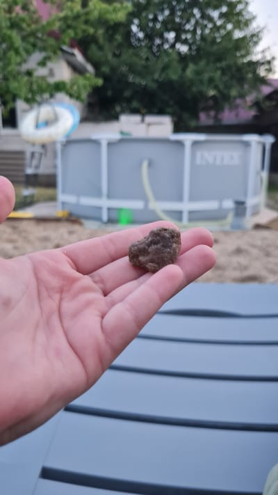 A person's hand is holding a small, dark, irregularly shaped rock. In the background, an above-ground swimming pool with the "INTEX" logo is visible. The scene appears to be an outdoor backyard setting, possibly with sandy ground. The location is Bilhorod-Dnistrovs'kyi, Ukraine.