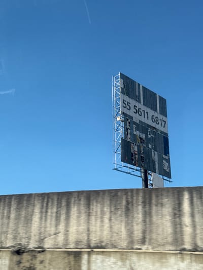 Looking up at a billboard from below, featuring a grid of panels. The number "55 5611 6817" is prominently displayed in white on a dark background. The billboard's structure is metal, appearing somewhat aged and weathered. The sky is clear and blue, and a vertical concrete structure partially obstructs the right side of the view.