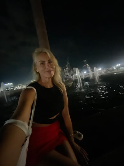 A woman with blonde hair is taking a selfie at night. She is wearing a black crop top and a red skirt. She is holding a white bag. In the background, there are fountains and buildings with lights. The scene is dark, suggesting it is nighttime. The location is Merchanskoe, Russia.