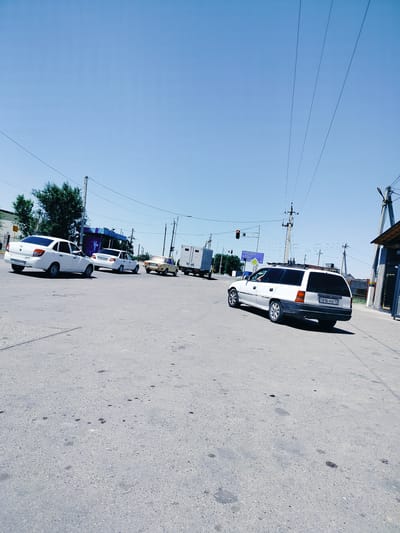The image depicts a bright, sunny day at a street intersection in Kölkent, Kazakhstan. Several vehicles are visible on the asphalt road. In the foreground and slightly to the right, a white station wagon is turning, with its rear end angled towards the viewer. To the left, a white sedan is parked or stopped. Further back, another white sedan is present, and behind it, a light-colored car is turning. A white truck with a trailer is also positioned at the intersection. Overhead power lines crisscross the clear blue sky, supported by utility poles. Trees line the background, and a bus stop shelter with a blue roof and purple supports is situated on the left side of the road. A traffic light is visible, displaying a red signal. The ground is paved with asphalt and shows some dark stains.