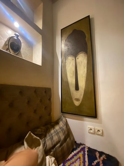 The image shows an interior scene in Taghazout, Morocco, featuring a bed with a tufted headboard and several pillows. On the wall, a large, framed abstract painting of a face with exaggerated features is displayed. To the left of the painting, a recessed shelf holds a decorative heart-shaped object. Two electrical outlets are visible on the wall below the painting. The lighting in the scene is dim, with a spotlight illuminating the shelf. The overall aesthetic appears to be bohemian or artistic.