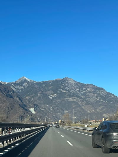 This image captures a bright, clear day along a multi-lane highway in a mountainous region, consistent with the landscape of Verrès, Italy. The prominent subject is a vast, rugged mountain range dominating the left side of the frame, showcasing dark rock faces and scattered lighter patches, possibly snow or lighter rock formations. The highway occupies the right side, extending into the distance, with distinct white lane markings and a metal guardrail running parallel to the mountain.
In the upper right foreground, a dark-colored car, possibly an SUV, is partially visible, indicating the photo was taken from a moving vehicle. Other vehicles appear as small dots in the distance on the highway. Along the base of the mountain, some indistinct buildings and infrastructure can be seen, suggesting a mix of natural landscape and human presence. The sky is a cloudless, vibrant blue, indicating fair weather and bright daylight. There is no discernible text visible in the image. The scene suggests travel through a scenic alpine environment.