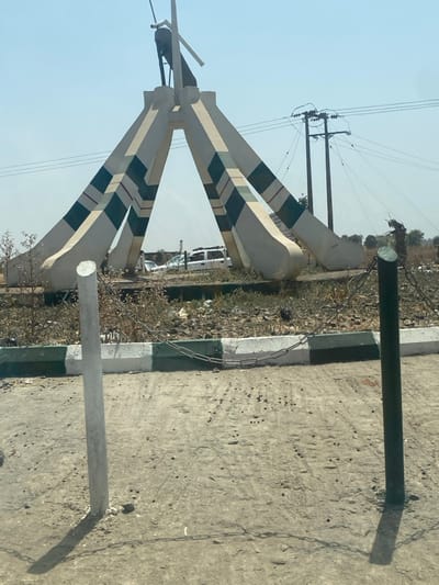 This image displays a street scene in Unguwar Gaya, Nigeria, under bright, clear daylight conditions. The central subject is a monument featuring four robust, upward-tapering concrete pillars painted white with horizontal green and thin red stripes, converging at an apex. A dark, abstract figure, possibly avian or mechanical, is mounted at the monument's peak.
In the immediate foreground, a light-colored, uneven dirt surface extends across the lower frame. A white vertical post is situated on the left, and a dark green/black vertical post is on the right. These posts are linked by a single strand of silver-colored metal chain, forming a low barrier. Behind this barrier, a concrete curb, painted with alternating white and dark green sections, defines the edge of a circular central island. This island's surface consists of dry, sparse vegetation, exposed dirt, and scattered debris.
In the background, behind the monument, a white SUV or van is partially visible, alongside another darker vehicle, suggesting a road or parking area. A solitary, indistinct figure is faintly discernible near the white SUV. To the right, multiple wooden utility poles with an array of overhead power lines are visible against a pale blue, cloudless sky. Distant, dry vegetation and minimal structures line the horizon.