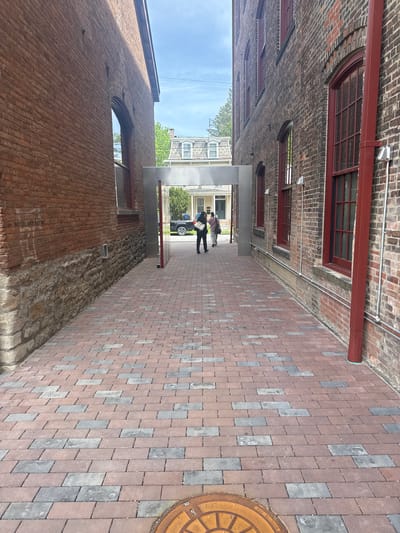 This media file captures an outdoor alleyway or narrow passage between brick buildings in the City of Hudson, United States of America. The ground is paved with rectangular reddish-brown and grey bricks. A circular, rusty-orange manhole cover is visible on the right side of the pavement, partially displaying the text "MADE IN USA".
The walls on both sides are constructed of brick, exhibiting varying textures and signs of age. The building on the right features several arched and rectangular windows with dark frames. A red-painted pipe or drainage system runs along the upper part of this building, accompanied by silver conduits. The building on the left also has brick walls, with a lower section appearing as rough stone masonry, and an arched window higher up.
At the far end of the alley, a modern, light-grey archway frames an opening. Two individuals are standing beneath this archway. One person wears a light-colored top and dark pants, while the other wears a dark top and dark pants. Both appear to be holding items. Beyond the archway, a bright blue sky with light clouds, distant buildings, green trees, and a dark car are visible. The scene suggests a clear, sunny daytime. No specific event or strong emotions are discernible; it's a casual urban street scene within what appears to be a historic district.