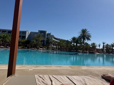 This is a wide-angle, eye-level outdoor shot of a resort pool area in Yasmine Hammamet, Tunisia. The foreground is dominated by a light brown chaise lounge with a beige towel draped over it, and a portion of a person's bare leg and foot is visible on the right. A large, reddish-brown wooden post is positioned vertically on the left side of the frame, partially obscuring the view.
Beyond the chaise lounge, a large, bright blue swimming pool stretches out, its surface shimmering with sunlight. The pool is bordered by a light-colored tiled walkway. In the middle ground, a row of thatched-roof cabanas with lounge chairs can be seen along the edge of the pool, with some guests relaxing in them. Palm trees are scattered throughout the scene, adding to the tropical ambiance.
In the background, a multi-story, grey resort building with balconies and windows curves along the horizon. The sky above is a clear, vibrant blue with a few distant birds. The overall scene conveys a sense of relaxation and leisure on a sunny day.