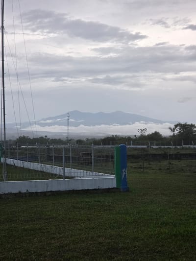 The image shows a landscape in Pintomakmur, Indonesia. A wire fence runs along the right side of the frame. Beyond the fence, there is a green, grassy area. In the distance, mountains are visible through a cloudy sky. A communications tower can be seen amidst the trees. Power lines are present in the foreground against the sky.