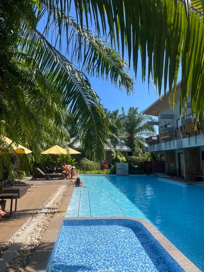 A sideways view captures a bright, sunny day at a tropical resort. A long, sparkling blue swimming pool stretches alongside a building with balconies. Lush green palm trees frame the scene, their fronds reaching towards a clear blue sky. Yellow umbrellas shade lounge chairs where people are relaxing near the pool's edge. The pool transitions into a shallower, tiled area, adding visual depth.
