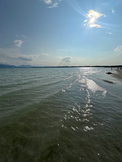 A landscape photograph captures a serene view of the Chiemsee in Germany. The foreground is dominated by clear, shallow water, revealing the sandy bottom beneath. Gentle ripples create a shimmering effect as sunlight reflects off the surface. A narrow strip of the shore runs horizontally across the image, with a few scattered people visible in the distance on the right side of the frame. The middle ground consists of the vast expanse of the lake, stretching towards a distant shoreline dotted with trees. In the background, a hazy mountain range forms a subtle silhouette against the horizon. The sky above is a vibrant blue, punctuated by scattered white clouds. A bright sun, partially obscured by a cloud, beams down, casting lens flares and accentuating the sunlit patterns on the water. The overall atmosphere is peaceful and summery.