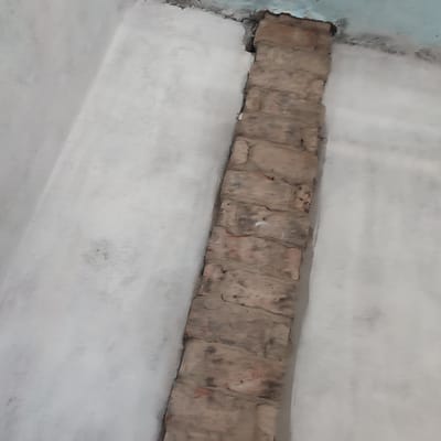 The image shows a close-up view of a wall where a vertical brick column protrudes from a plastered surface. The brickwork appears old and weathered, with visible mortar lines and cracks. The plastered wall is painted a light, neutral color and shows some minor imperfections and discoloration. The scene is indoors, likely in a building under construction or renovation, as suggested by the raw brickwork and unfinished plaster. The lighting is somewhat dim, casting subtle shadows that highlight the textures of the materials. There are no people or discernible actions occurring in the frame. The location of Alga, Kyrgyzstan is not visible in the image.