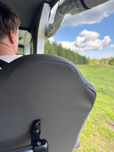 The media file shows a close-up of a dark gray textured seat, likely inside a vehicle or boat, with a person's head and shoulder visible in the lower left. The person appears to be sleeping or resting, with their eyes closed and head tilted back. The seat has a stitched seam running along its curve.

Through a window or opening to the left of the seat, a bright blue sky with fluffy white clouds is visible, suggesting it is daytime. The background beyond the seat is a blur of green, indicating an outdoor natural setting, possibly a field or grassy area with trees in the distance. The overall impression is of a relaxed moment during a journey on a pleasant day.

The scene is set in Nechkino, Russia. There is no visible text in the image. The lighting suggests it is daytime, and the clear sky implies fair weather. The focus on the seat and the sleeping person suggests a moment of downtime or travel.