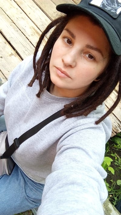 A medium shot of a fair-skinned person with brown dreadlocks, wearing a gray sweatshirt, jeans, and a black baseball cap. The person has a piercing in their nostril. A black strap crosses their chest. The background consists of wooden planks and some green foliage. The location is potentially in Юдино, Russia, though this cannot be definitively confirmed from the image alone.
