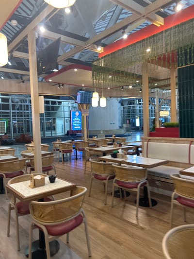 The image depicts the interior of a restaurant or cafe. The decor features natural wood elements, including structural beams and table legs, complemented by woven rattan chairs with red upholstered seats. The tables have light-colored, patterned tops. In the background, a large window or glass wall offers a view of an airport concourse, indicated by the presence of seating areas, signs, and the general architecture of a travel hub. Hanging plants and green accent features add a touch of biophilic design. The lighting is a mix of ceiling lights and pendant lamps. The scene is clean and orderly, with no customers or staff immediately visible. The location is Baji Mangngai, Indonesia, likely within the airport facilities.