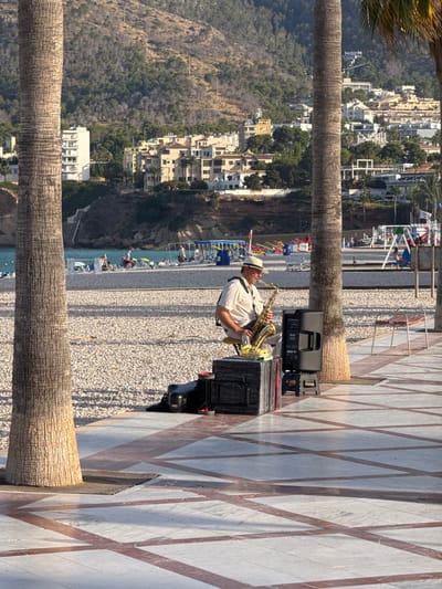 An adult male wearing a white hat and a light-colored shirt is playing a saxophone on a pebbled beach. He is seated on a small stool next to a black speaker and a black case. In the background, there are palm trees, buildings on a hillside, the sea, and other beachgoers relaxing on lounge chairs. The foreground shows a patterned tiled walkway. The scene is set in l'Alfàs del Pi, Spain, specifically on the beach.