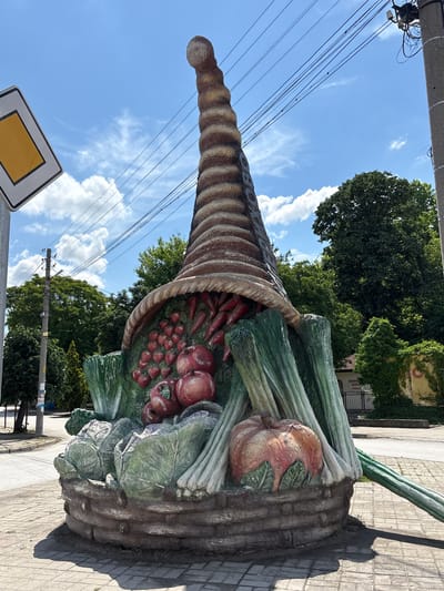 The image shows a large, sculpted cornucopia, or horn of plenty, overflowing with vegetables, including tomatoes, onions, leeks, and cabbages. The sculpture is located in the city of Biaga, Bulgaria. The cornucopia is made of a textured, weathered material, likely concrete or similar, and painted to resemble woven straw and the colors of the various vegetables. It is situated on a paved walkway near a street with overhead power lines and foliage. In the background, there are trees and possibly a building. A yellow diamond traffic sign is visible near the bottom left corner of the image. The sky is blue with scattered clouds.