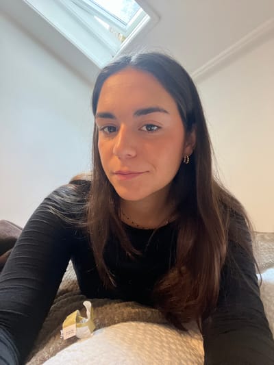 The image displays a young adult female with long, dark brown hair, wearing a black long-sleeved top and small gold hoop earrings, looking directly at the camera with a subtle smile. She is positioned in a reclining manner on light-colored, textured bedding or upholstery. To her lower left, a crumpled, yellowish-white wrapper with a tag bearing the number "713172" is visible. The background features a plain, light-colored wall and a partially open skylight window in the upper left, revealing outdoor foliage. The specific location within Bath, United Kingdom, cannot be determined from the visual evidence.