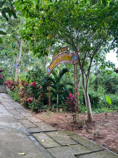 The image shows a paved pathway leading to an archway with a sign in front of it. The sign reads "SELAMAT DATANG TAMAN PKK Ds. KARANGLOR" in yellow lettering on a brown background. The pathway is made of gray concrete blocks and is surrounded by lush green vegetation. The archway is constructed of metal and supports a brown sign. The sign is supported by two upright posts connected by a horizontal beam. The area is located at coordinates -7.826, 110.816 in Kecamatan Manyaran, Indonesia. There are no people in the scene.