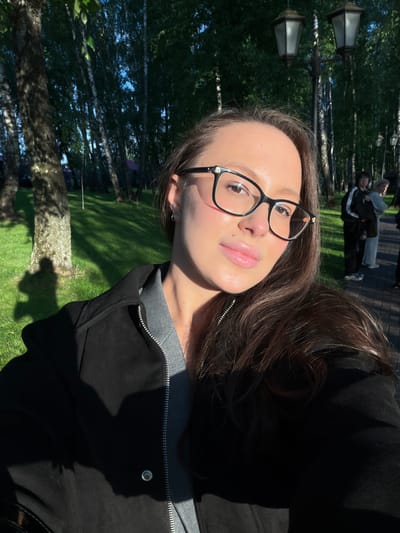 A young woman with long brown hair, wearing black-framed glasses, a gray shirt, and a black jacket, is shown in a selfie taken outdoors. The background reveals a park-like setting with birch trees and streetlights. In the distance, two people are seen lying on a paved surface. The photograph appears to be taken in Polivanovo, Russia, as indicated by the visual elements of the environment.