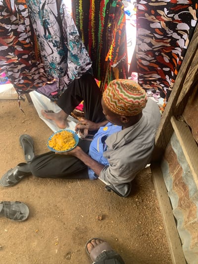 The image depicts a person sitting on the ground in what appears to be an outdoor setting in Project, Nigeria. The individual is wearing a grey hooded sweatshirt, a blue shirt underneath, black pants, and a multi-colored kufi cap. They are holding a blue plate filled with yellow food, identified as rice, and are in the process of eating it with a fork. The person is barefoot, wearing black sandals on their left foot, and other sandals are visible on the ground nearby, along with a second person's foot wearing brown sandals. The background features colorful fabrics hanging, suggesting a market or a domestic setting. The ground is composed of dirt and small stones. Wooden planks and corrugated metal are visible on the right side of the frame, indicating a rustic structure. The lighting suggests it is daytime.