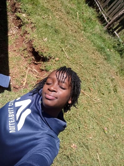 A single individual is depicted lying on their back outdoors in Waseges ward, Kenya, with the camera positioned directly above, capturing a selfie. The person, a young individual with dark skin and short, dark braids partially covering the forehead, wears a dark blue long-sleeved hooded sweatshirt featuring a white, mirrored graphic on the left chest ("AOTTELRVT C" partially legible). Their eyes are closed, and lips are slightly parted in a relaxed expression. The background consists of green grass with visible patches of reddish-brown soil and scattered dried plant material. A dark wooden fence is visible in the upper right background, and sunlight illuminates the scene, casting a dark shadow in the upper left corner, where a piece of light blue fabric is also partially visible.